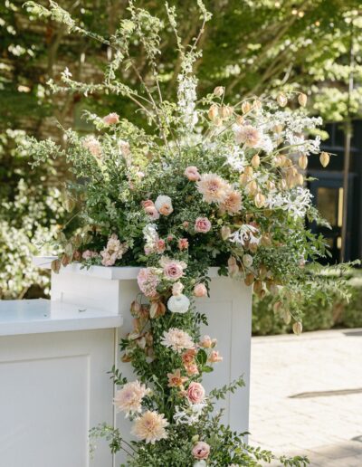 Floral arrangement with pink and white flowers and green foliage on a white stand outdoors.