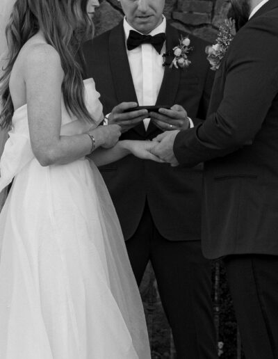 A bride and groom hold hands during their wedding ceremony, with an officiant reading from a device. They stand in front of a stone wall.