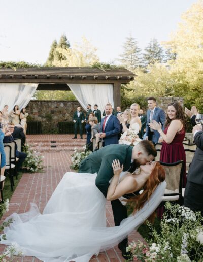 A groom kisses his bride, dipping her backward, in an outdoor wedding ceremony. Bridesmaids, groomsmen, and guests stand around, smiling and clapping.