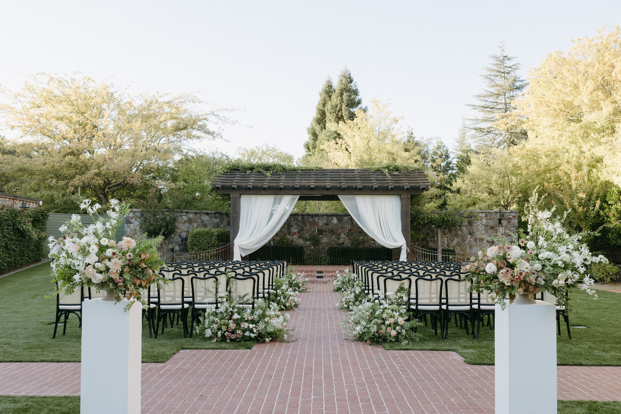 Outdoor wedding ceremony setup with chairs facing an altar, draped with white curtains. Expertly arranged by an Estate Yountville Wedding Planner, the floral arrangements on pillars and along the aisle are stunningly elegant, all surrounded by serene trees and lush greenery.