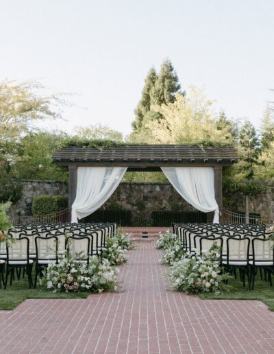 Outdoor wedding ceremony setup with rows of black chairs facing a pergola draped in white fabric, surrounded by greenery and floral arrangements on a brick pathway.