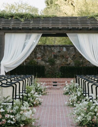 Outdoor wedding ceremony setup with rows of black chairs facing a pergola draped in white fabric, surrounded by greenery and floral arrangements on a brick pathway.