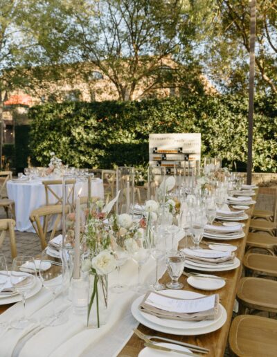 Elegant outdoor dining setup with a long wooden table, white floral centerpieces, candles, and neatly arranged tableware. Surrounded by greenery and wooden chairs.