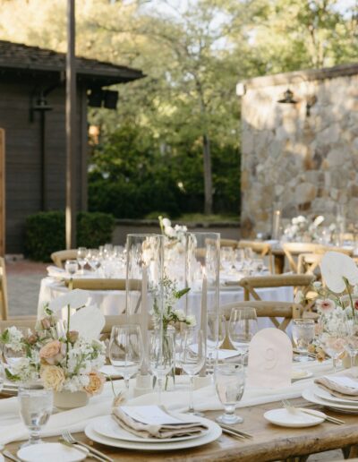 Outdoor wedding reception setup with rustic wooden tables, elegant white and beige floral arrangements, white plates, and tall glass candle holders against a stone wall backdrop.