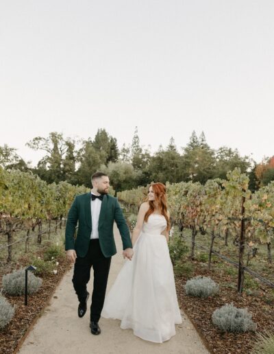 A couple in formal attire walks hand in hand along a path in a vineyard.