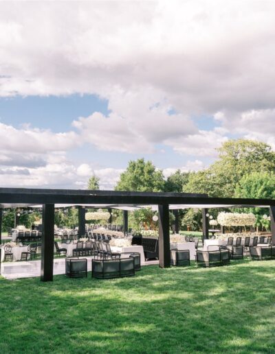 Outdoor event setup with black wooden canopy, chairs, and tables on a grassy area, surrounded by trees under a partly cloudy sky.