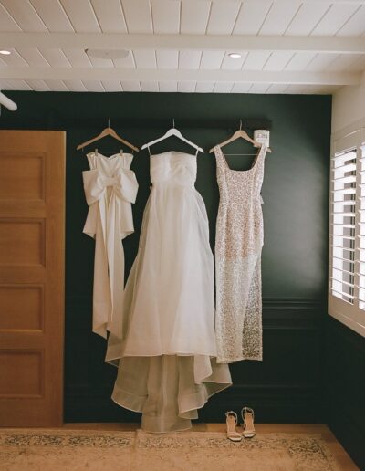 Three dresses, including a strapless gown and a lace dress, hang on a dark wall with a pair of sandals below.