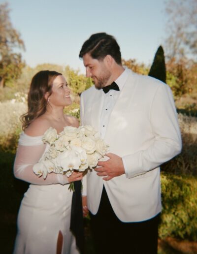 A couple dressed in formal attire stands outdoors, smiling at each other. The woman holds a bouquet of white flowers.