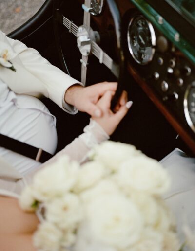 A couple holds hands inside a vintage car, with a bouquet of white flowers in the foreground.