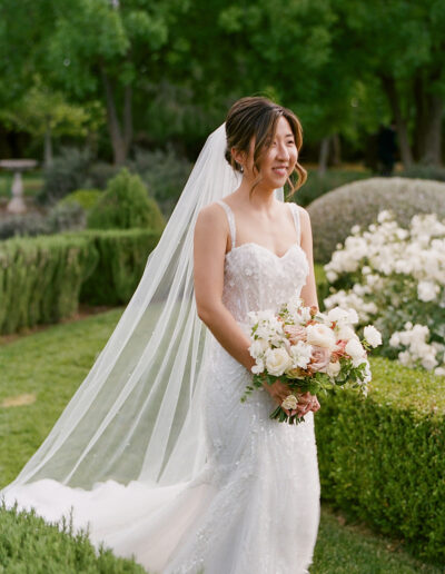 The bride, in a white dress with a delicate veil, holds a bouquet of flowers while standing amidst the enchanting greenery and white blooms of Park Winters Weddings.