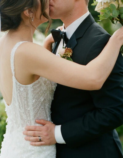 At a Park Winters wedding, a couple in formal attire shares a tender kiss. The woman stuns in a white beaded dress, while the man complements her look with his black suit and floral boutonniere.