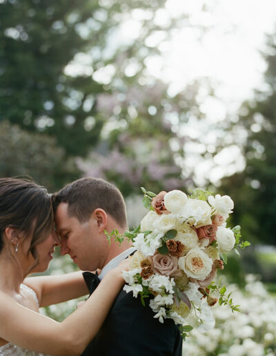 A couple stands closely, touching foreheads at a Park Winters wedding, with a bouquet of white and blush flowers in the foreground, surrounded by greenery and soft lighting.