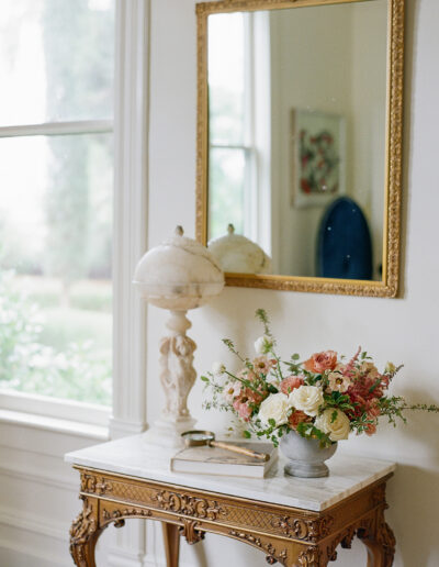 A decorative table with a marble top graces the room, holding a floral arrangement reminiscent of Park Winters Weddings, alongside a small globe lamp and a book. Above it, an ornate gold mirror captures the elegance of such memorable occasions.