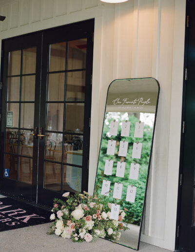 A mirror seating chart with white cards numbered 1 to 13 is beautifully adorned with a flower arrangement at the base, leaning elegantly against the closed glass-paneled doors, capturing the enchanting essence of Park Winters weddings.