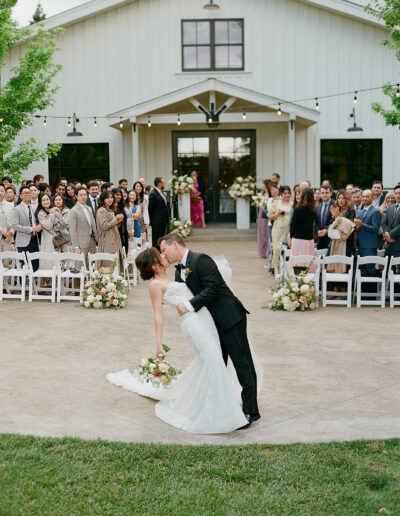 At a Park Winters wedding, a bride and groom share a kiss during their ceremony, surrounded by guests seated in white chairs. The romantic event unfolds outdoors, with the elegant white building providing a stunning backdrop.