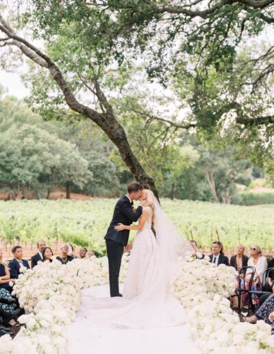 A bride and groom kiss at an outdoor wedding ceremony, surrounded by guests and white flowers, with a vineyard in the background.