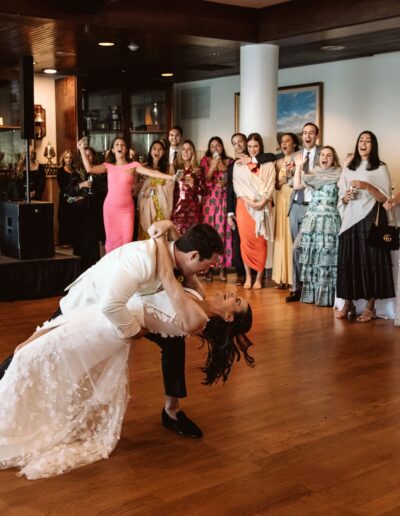 A couple in wedding attire shares a dance while guests watch in a reception hall. A band is set up onstage in the background.