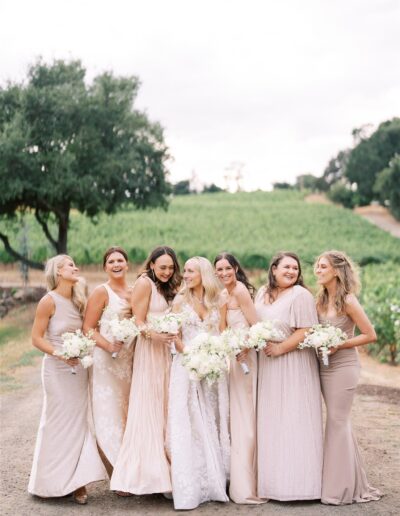 A bride stands with six bridesmaids in pastel dresses, holding bouquets, on a path with greenery and trees in the background.