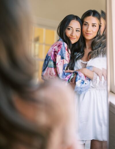 Two women stand in front of a mirror. One woman is wearing a floral robe, and the other is in a white outfit. They are gazing at their reflection together.