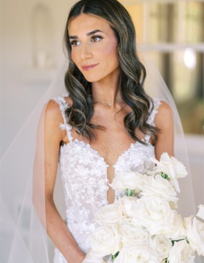 Bride in a lace wedding dress holds a bouquet of white roses, standing indoors with a soft background.