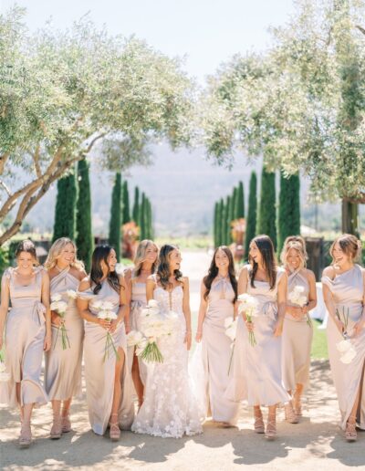 A bride in a white dress stands with seven bridesmaids in light dresses, holding white flowers, outdoors under a clear sky with trees lining the path.