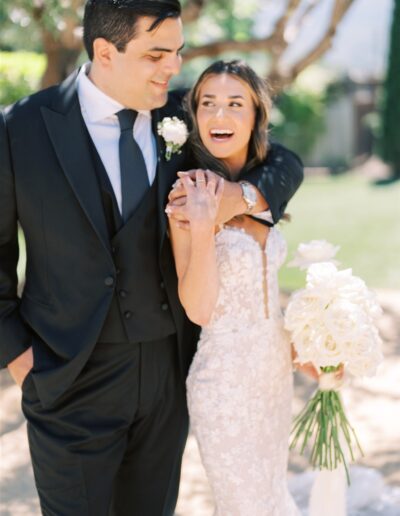 A bride and groom stand outdoors, smiling. The bride holds a bouquet of white flowers while the groom has his arm around her shoulder. They are in formal wedding attire.