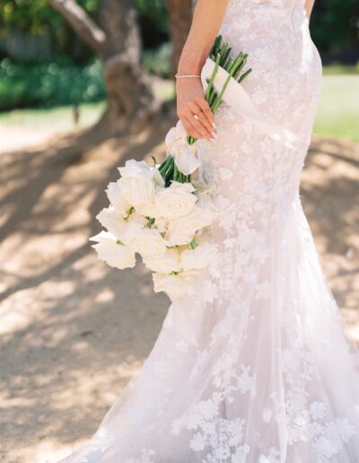 Bride in a lace gown holding a bouquet of white roses outdoors.