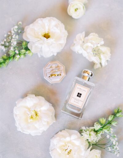 Perfume bottle surrounded by white flowers and a velvet ring box with two rings, placed on a light gray surface.