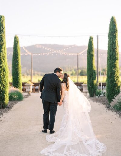 A couple in wedding attire walks along a path, holding hands, surrounded by tall trees and lights.