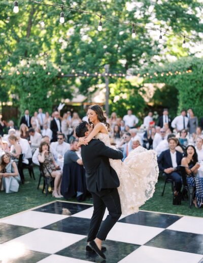 A bride and groom dance on a checkered floor outdoors, surrounded by seated guests. String lights hang above, and trees provide a green backdrop.