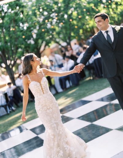 A couple dances together on a black and white checkered floor outdoors, surrounded by guests and greenery.