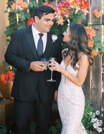 A couple in formal attire stand outdoors, holding wine glasses. The man wears a black suit, and the woman wears a white lace dress. They are surrounded by vibrant flowers.