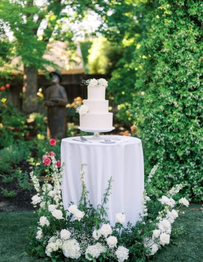 A white three-tiered cake on a pedestal table, surrounded by white and green floral arrangements in a garden setting.