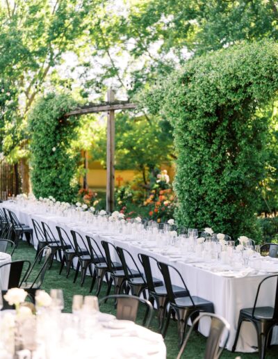 Long tables with white tablecloths and black chairs set in an outdoor garden with lush greenery and flowers.