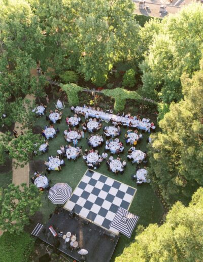 Aerial view of an outdoor event with round tables set for dining, a large checkerboard dance floor, and surrounding trees in a garden setting.