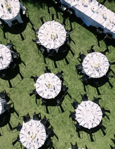 An aerial view of multiple round tables set for an outdoor event on a grassy area, with white tablecloths, black chairs, and floral centerpieces.