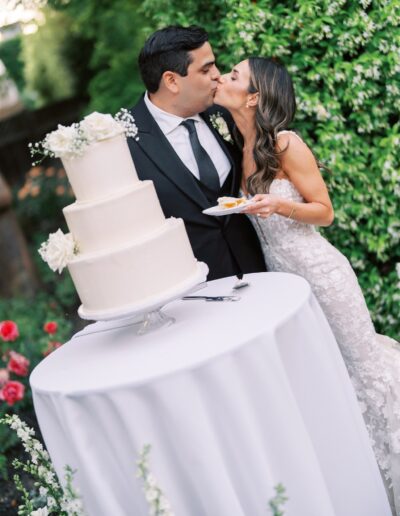 A couple kisses in front of a three-tiered white wedding cake on a table outdoors. The bride holds a plate with a slice of cake.