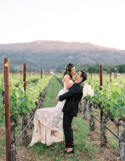 A groom lifts a bride in a vineyard, both smiling, with a mountain in the background.