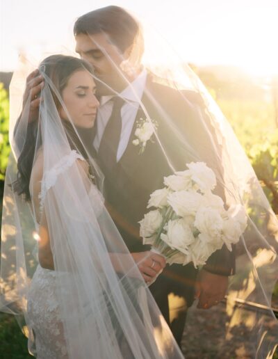 A bride and groom stand under a sheer veil in an outdoor setting. The bride holds a bouquet of white roses.