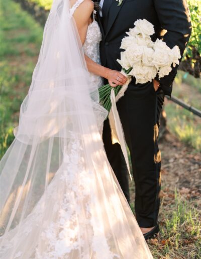 Bride and groom in a vineyard, with the bride wearing a lace gown and holding a bouquet of white roses.