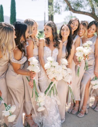 A bride in a white gown stands with bridesmaids in taupe dresses, all holding white rose bouquets, outdoors.