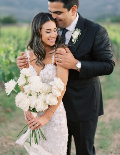 A couple in formal attire poses outdoors. The woman holds a bouquet of white roses, and the man embraces her from behind. They are in a natural setting with greenery in the background.