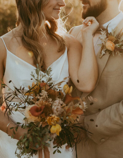 A couple stands together in a field at sunset, with the woman holding a bouquet of flowers. Both wear formal attire with floral accents.