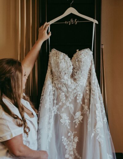 A person holds up a lace wedding dress on a hanger labeled "mrs," showcasing the detailed floral patterns on the gown.