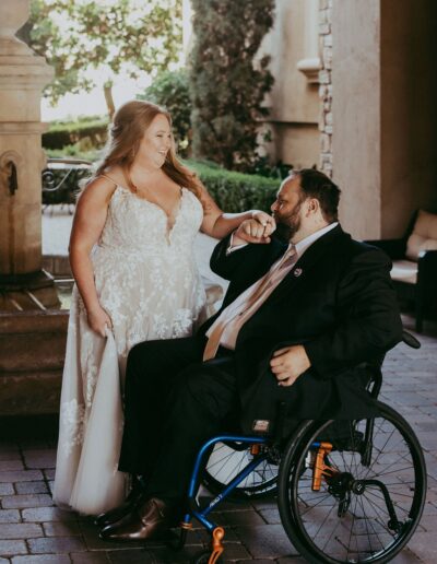 A bride in a white dress and a groom in a suit, seated in a wheelchair, share a joyful moment outdoors near a fountain.