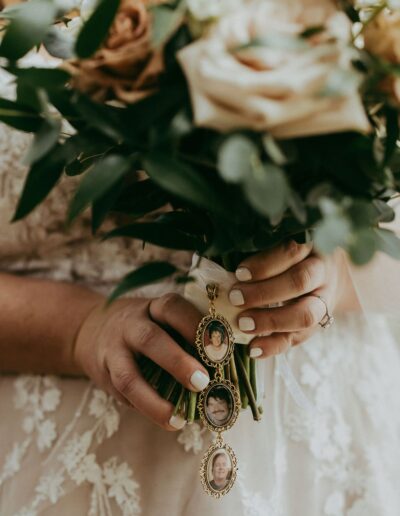 Close-up of a bride holding a floral bouquet with three photo lockets hanging, each displaying a portrait. The bride wears a lace dress and has painted nails.