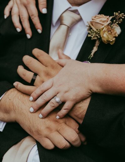 Close-up of a couple's hands with wedding rings, resting on the groom's chest. The groom wears a dark suit and beige tie, with a boutonniere featuring a peach rose.