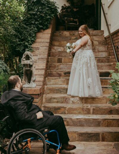 A woman in a wedding dress stands at the top of a stone staircase, looking back at a man in a wheelchair at the bottom, with greenery surrounding them.