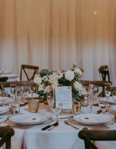 A wedding reception table with elegant place settings, floral centerpiece, candles, and a menu. In the background, similarly set tables are visible.