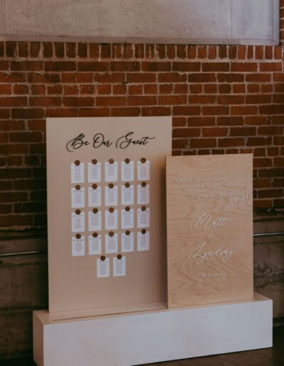 Two beige display boards stand against a brick wall. The left has organized cards labeled "Be Our Guest," and the right features a romantic quote with names and a date.